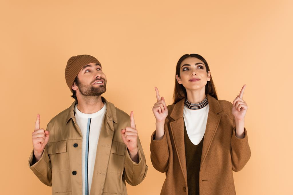 cheerful couple in stylish coat and jacket pointing up with fingers isolated on beige - Foto, immagini
