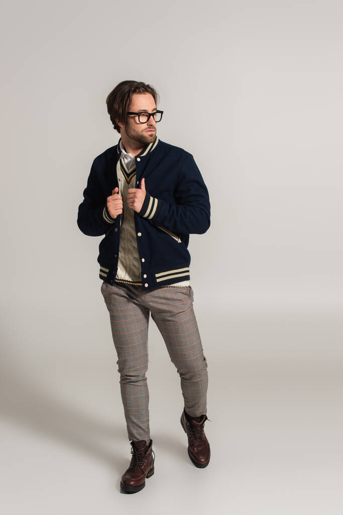 Вид в полный рост человека в стильной куртке, брюках и сапогах, отводящего взгляд на сером фоне - Фото, изображение