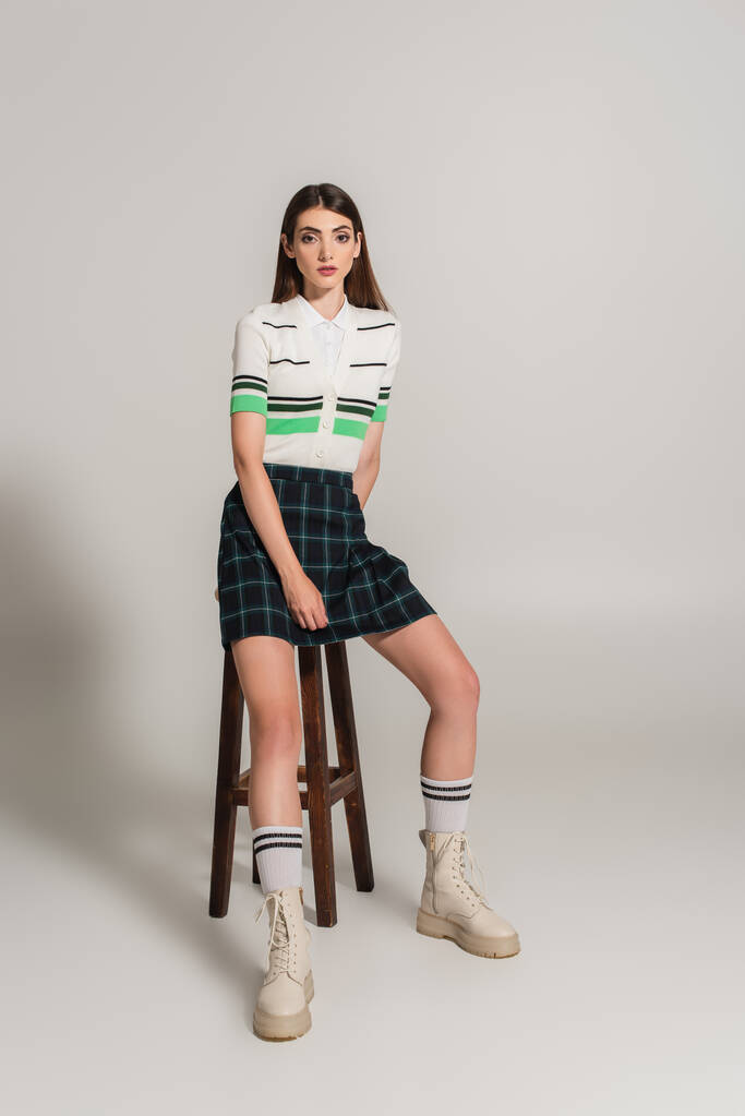 young woman in trendy clothes sitting on high wooden stool on grey background - Φωτογραφία, εικόνα