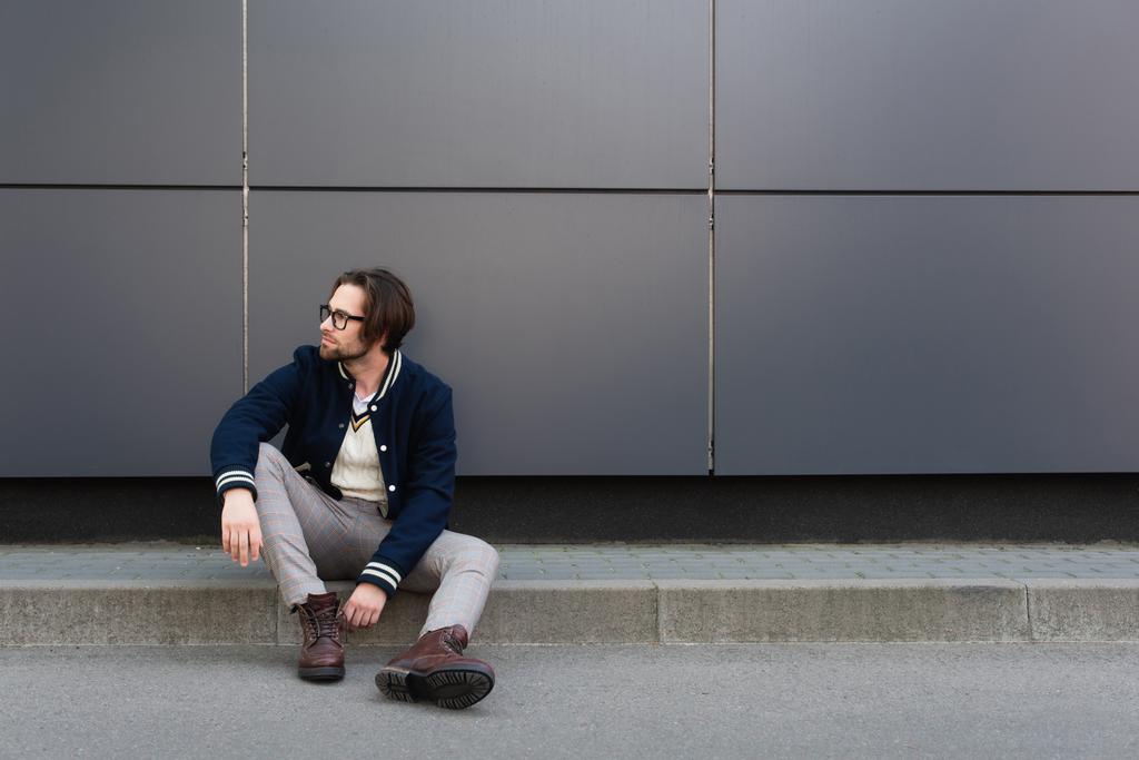 jonge man in trendy herfstkleding die wegkijkt terwijl hij op de rand bij de grijze muur zit - Foto, afbeelding