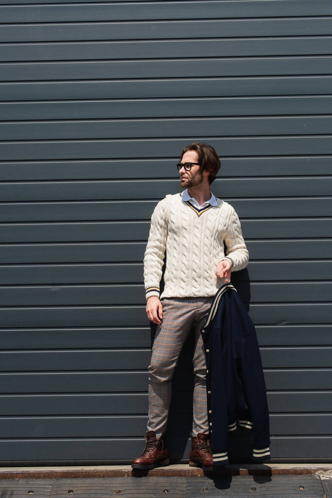 trendy man in white pullover holding blue jacket near grey wall outdoors - Fotografie, Obrázek