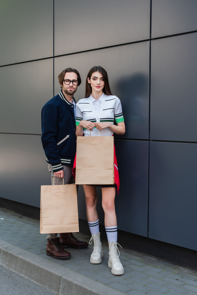 fashionable couple looking at camera while standing near grey wall with shopping bags - Photo, image