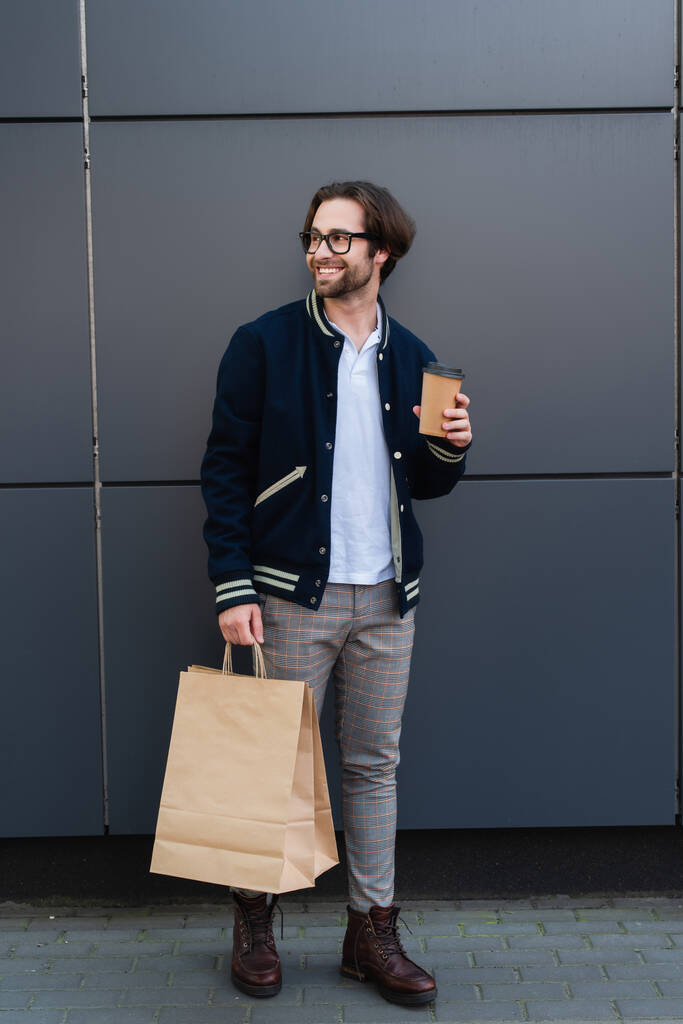 веселый и стильный мужчина, стоящий у серой стены с бумажной чашкой и пакетами для покупок - Фото, изображение