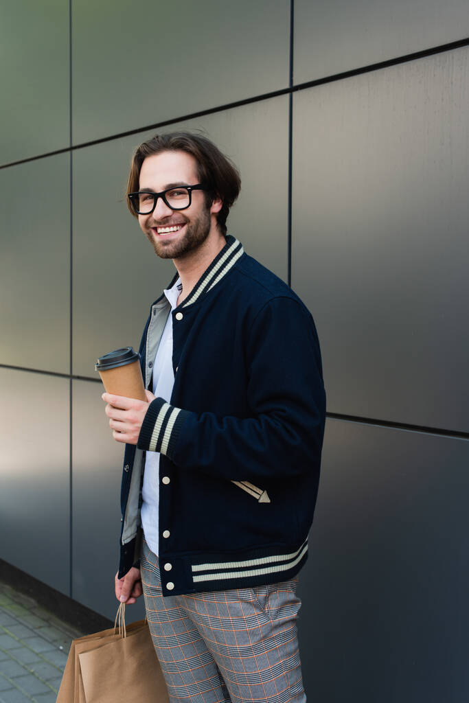 joyful man in trendy jacket and eyeglasses holding takeaway drink near grey wall - Foto, imagen