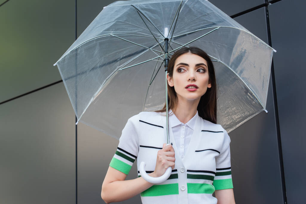 trendy vrouw staat onder transparante paraplu en kijkt weg - Foto, afbeelding