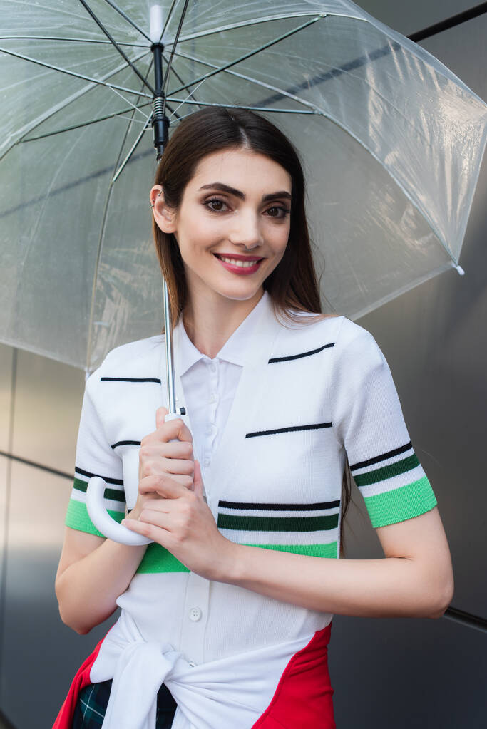 joyful woman in striped polo t-shirt smiling at camera under transparent umbrella - Photo, Image