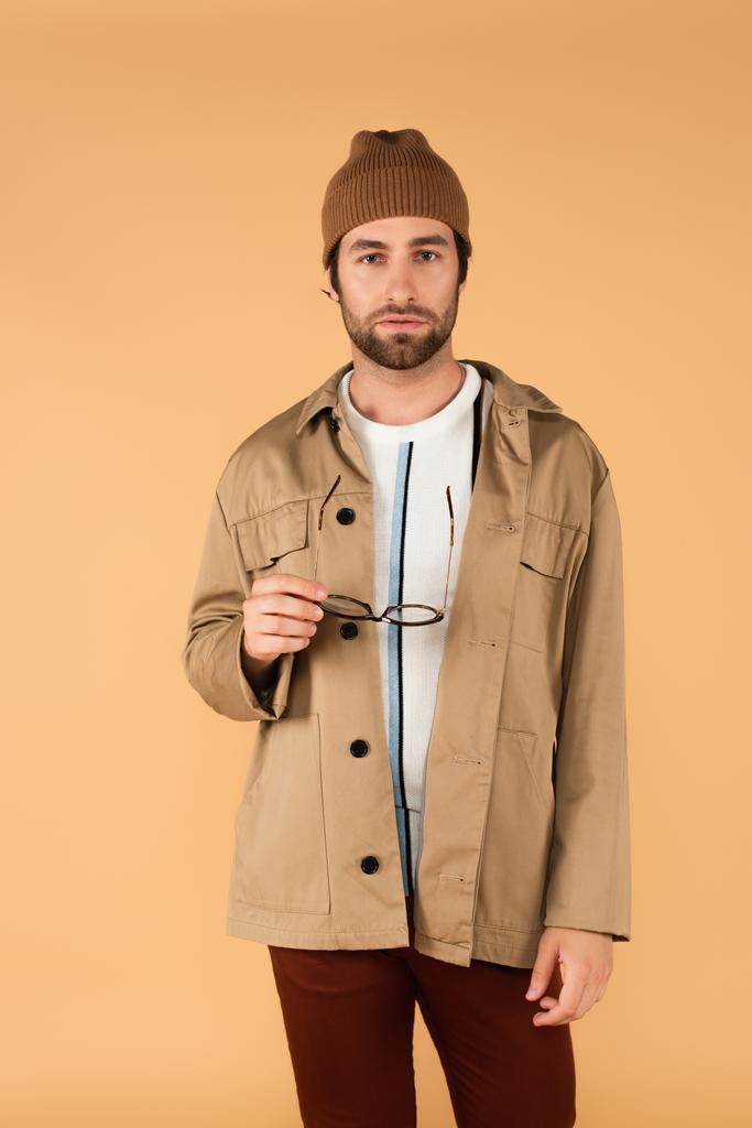 Вид спереди на молодого человека в стильной куртке и шапочке, держащего очки изолированные на бежевом - Фото, изображение
