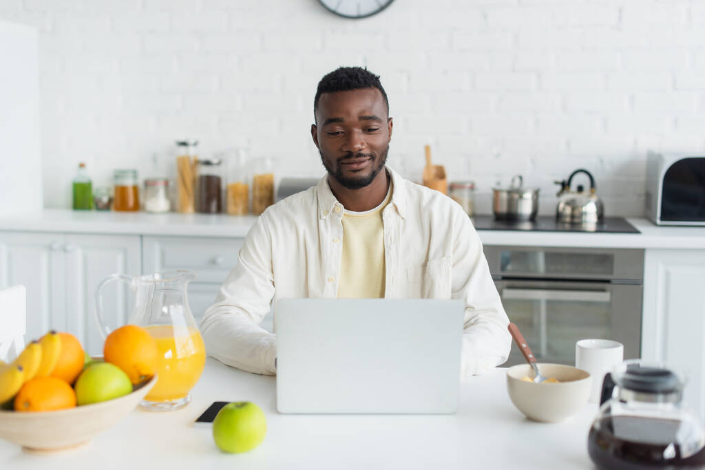 счастливый молодой афроамериканец использует ноутбук на кухне  - Фото, изображение