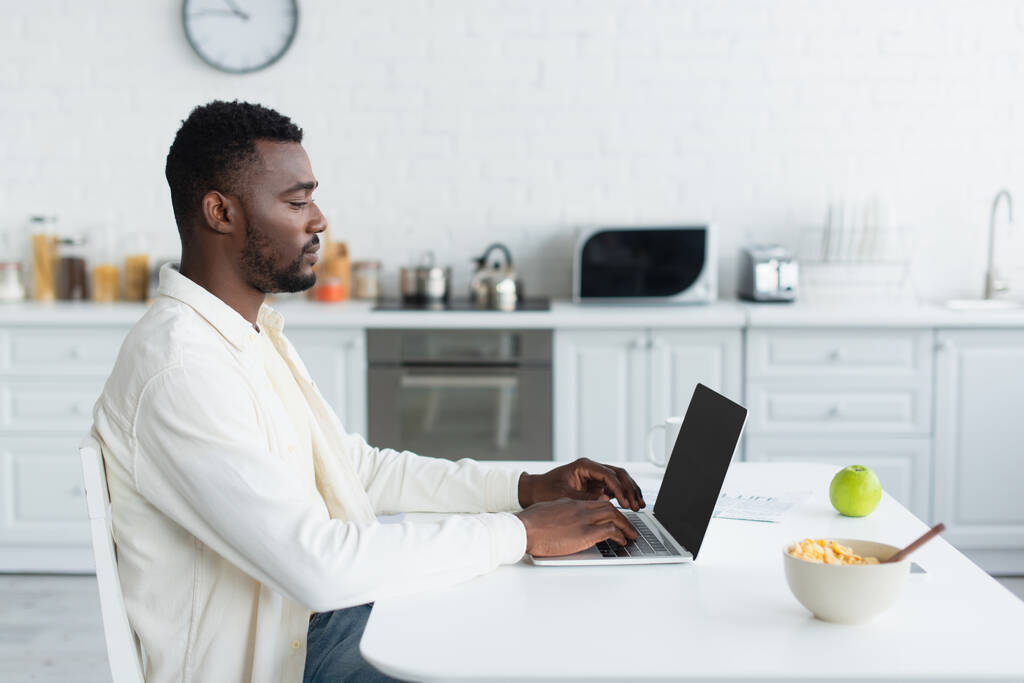 πλαϊνή άποψη του νεαρού αφροαμερικανού ελεύθερου επαγγελματία χρησιμοποιώντας φορητό υπολογιστή με λευκή οθόνη στην κουζίνα  - Φωτογραφία, εικόνα