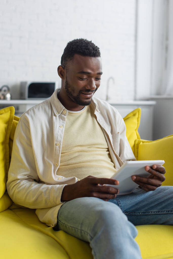 homme afro-américain joyeux utilisant une tablette numérique tout en étant assis sur le canapé  - Photo, image