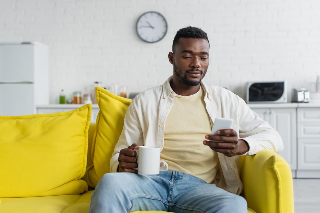 junger afrikanisch-amerikanischer Mann benutzt Smartphone, während er auf Couch mit Tasse sitzt - Foto, Bild