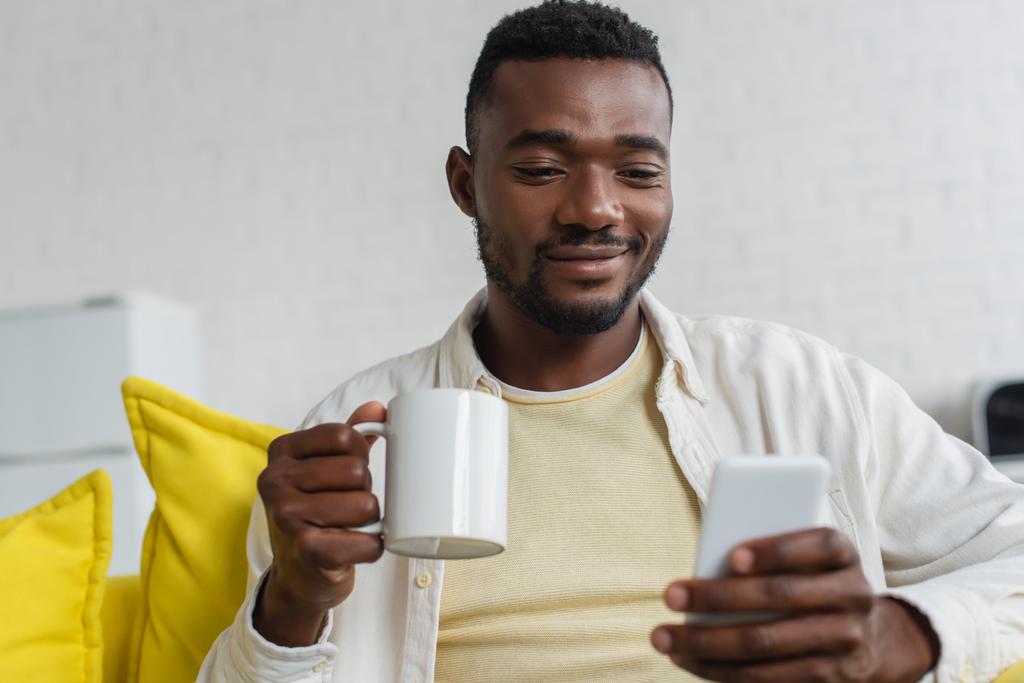 усміхнений молодий афроамериканський чоловік використовує мобільний телефон і тримає чашку
  - Фото, зображення
