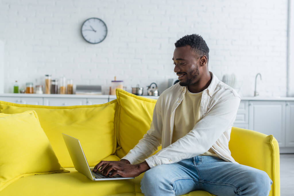 黄色のソファに座っている間にノートパソコンでアフリカ系アメリカ人のフリーランスが - 写真・画像