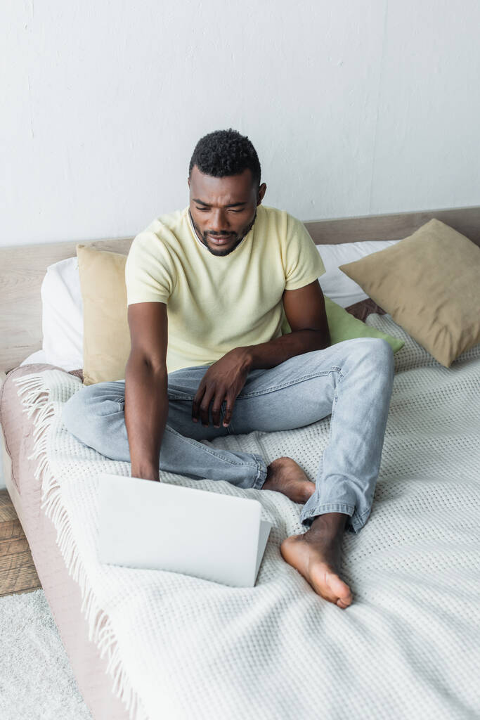 homme afro-américain pieds nus tapant sur ordinateur portable dans la chambre  - Photo, image