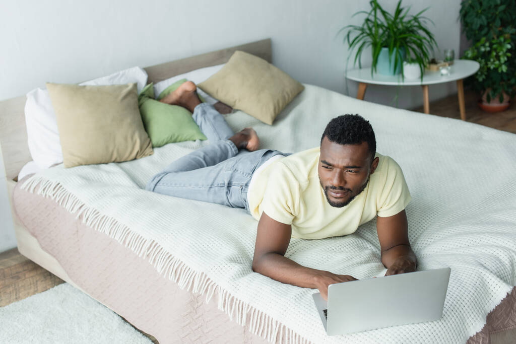 přemýšlivý afroameričan na volné noze v tričku pomocí notebooku a ležící na posteli - Fotografie, Obrázek