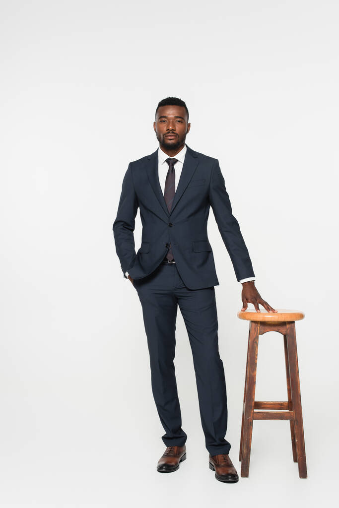 full length of african american businessman in suit posing with hand in pocket near stool isolated on grey - Photo, Image