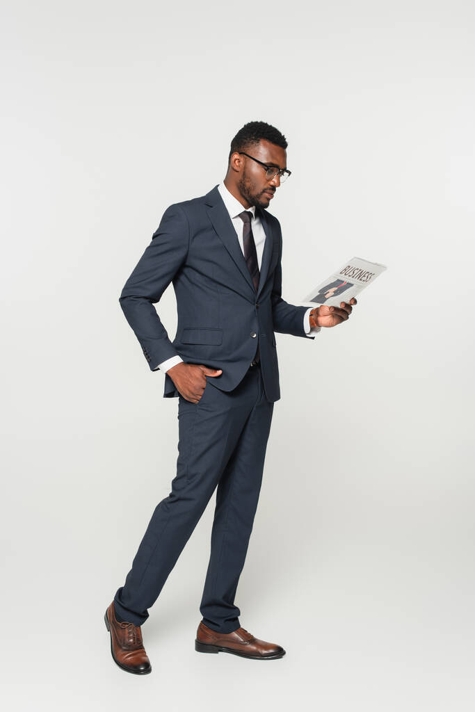intera lunghezza dell'uomo afroamericano negli occhiali da vista che legge il giornale di affari mentre si leva in piedi con la mano nella tasca isolata su grigio - Foto, immagini