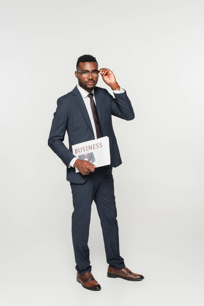 volledige lengte van de Afrikaans-Amerikaanse man aanpassen bril en bedrijf krant geïsoleerd op grijs - Foto, afbeelding