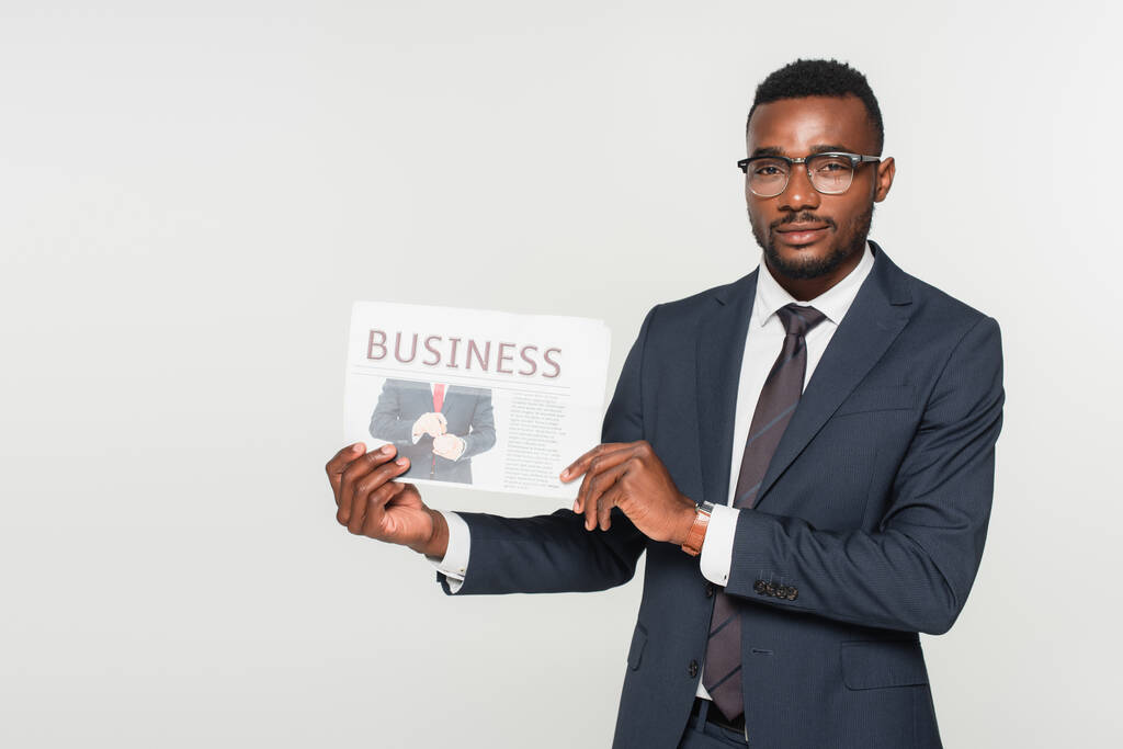 Afrikanisch-amerikanischer Mann mit Brille zeigt Wirtschaftszeitung isoliert auf grau - Foto, Bild