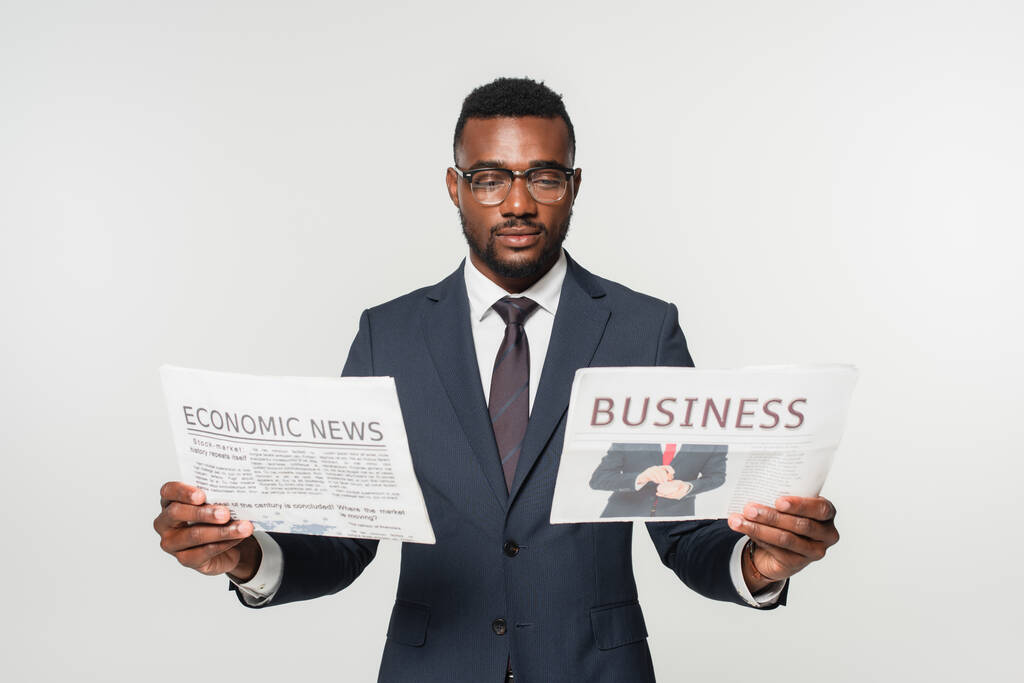 hombre afroamericano en gafas con periódicos con letras de noticias económicas y de negocios aisladas en gris - Foto, Imagen