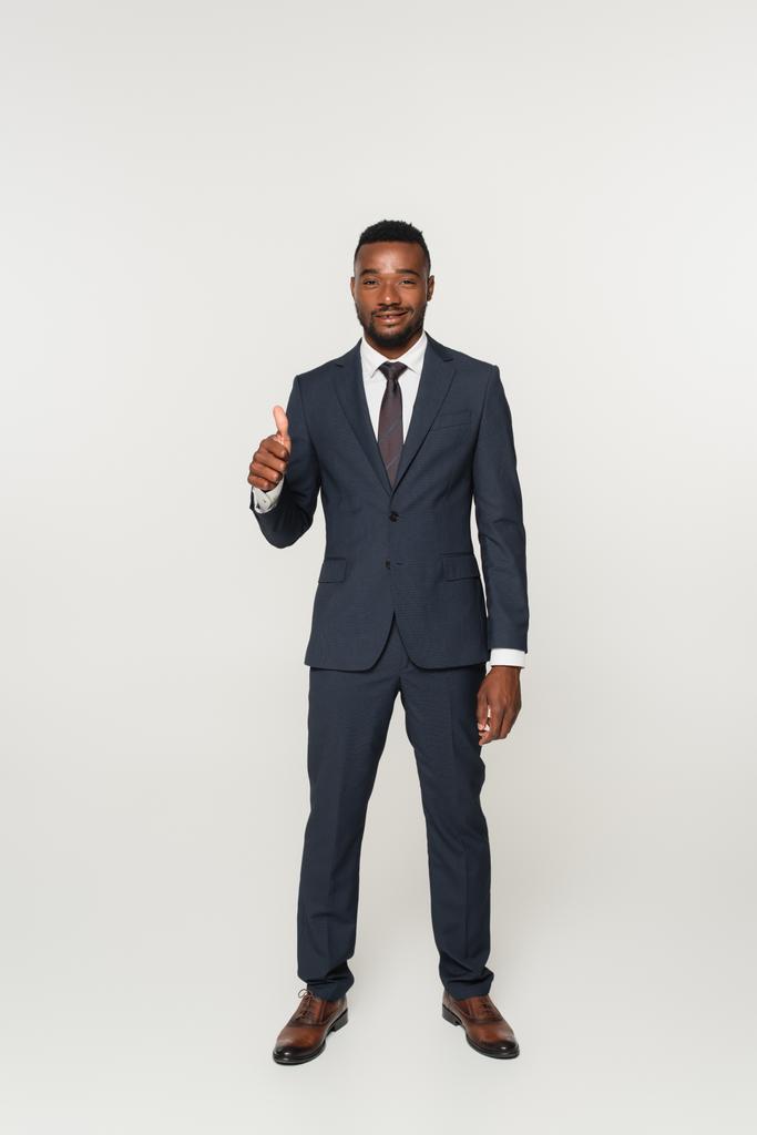 volle Länge des lächelnden afrikanisch-amerikanischen Geschäftsmannes im Anzug, der vereinzelt den Daumen nach oben auf grau zeigt - Foto, Bild
