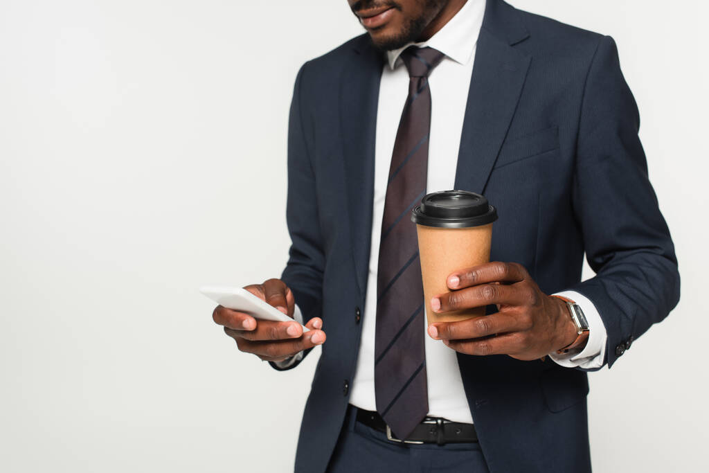 обрезанный вид на африканского американского бизнесмена в костюме обмена сообщениями на смартфоне и проведение бумажной чашки изолированы на серый  - Фото, изображение