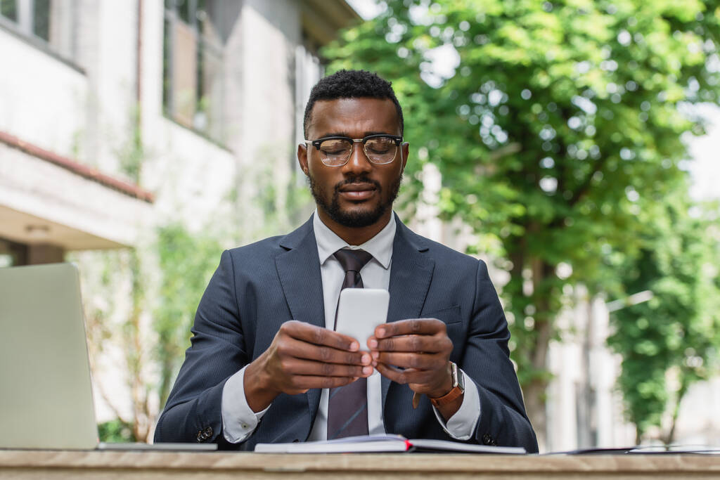 bebaarde Afrikaanse Amerikaanse zakenman in bril met behulp van smartphone in de buurt laptop - Foto, afbeelding