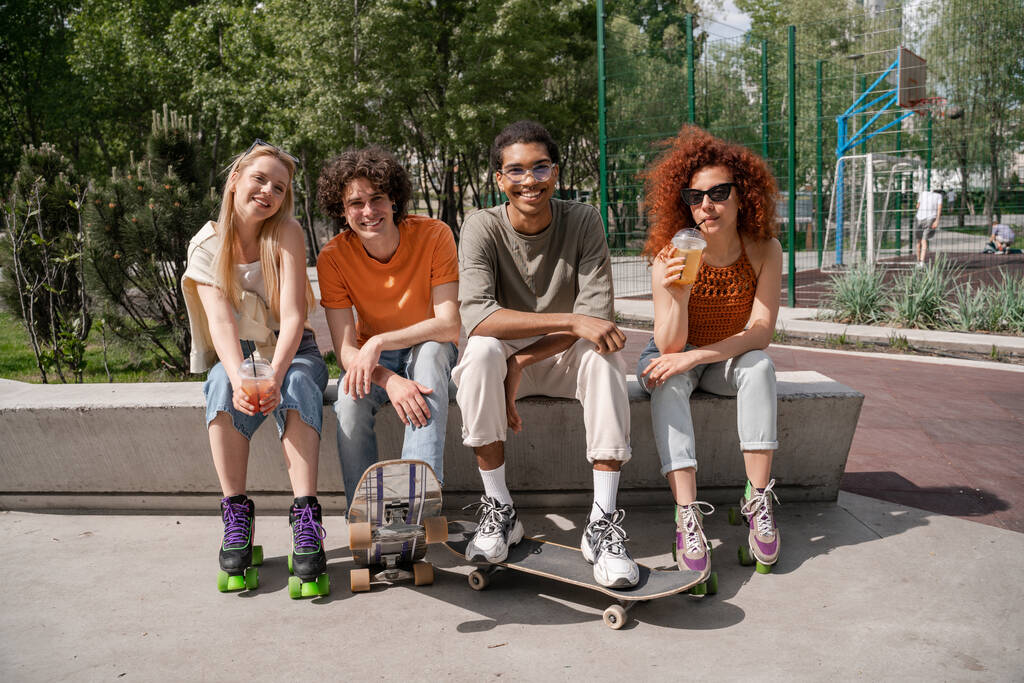 jeunes femmes heureuses avec des tasses en plastique assis à la frontière avec des patineurs interracial - Photo, image