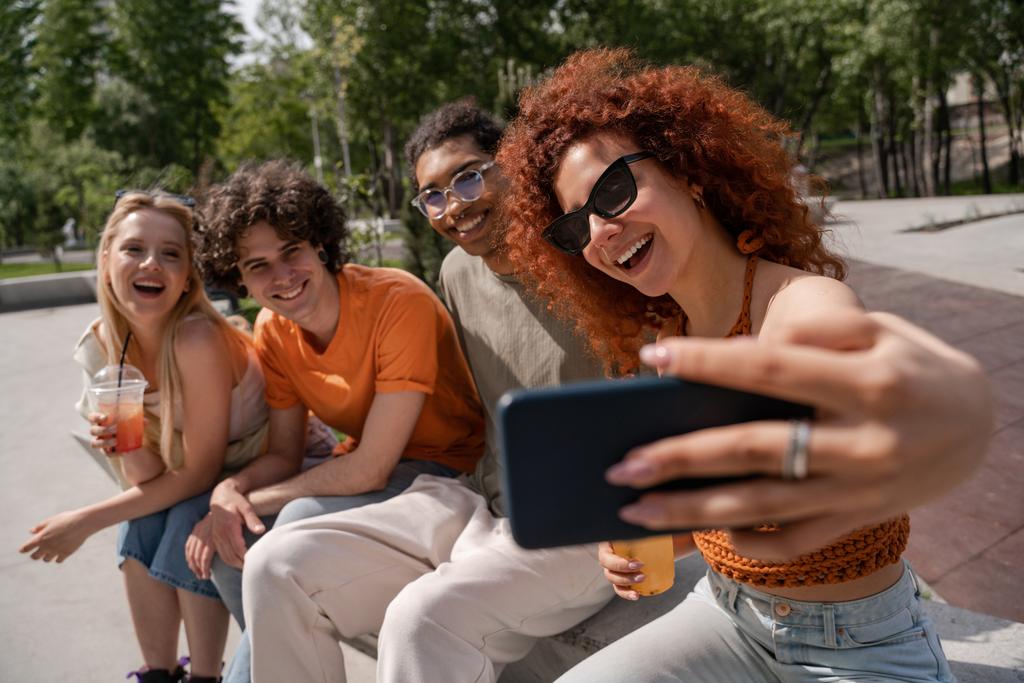 mulher encaracolado e alegre em óculos de sol tomando selfie com amigos multiétnicos - Foto, Imagem