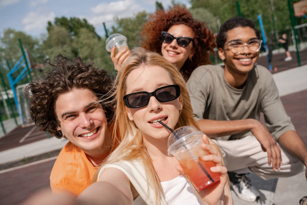 mujer joven en gafas de sol bebiendo refrescante cóctel cerca de amigos multiétnicos felices - Foto, imagen