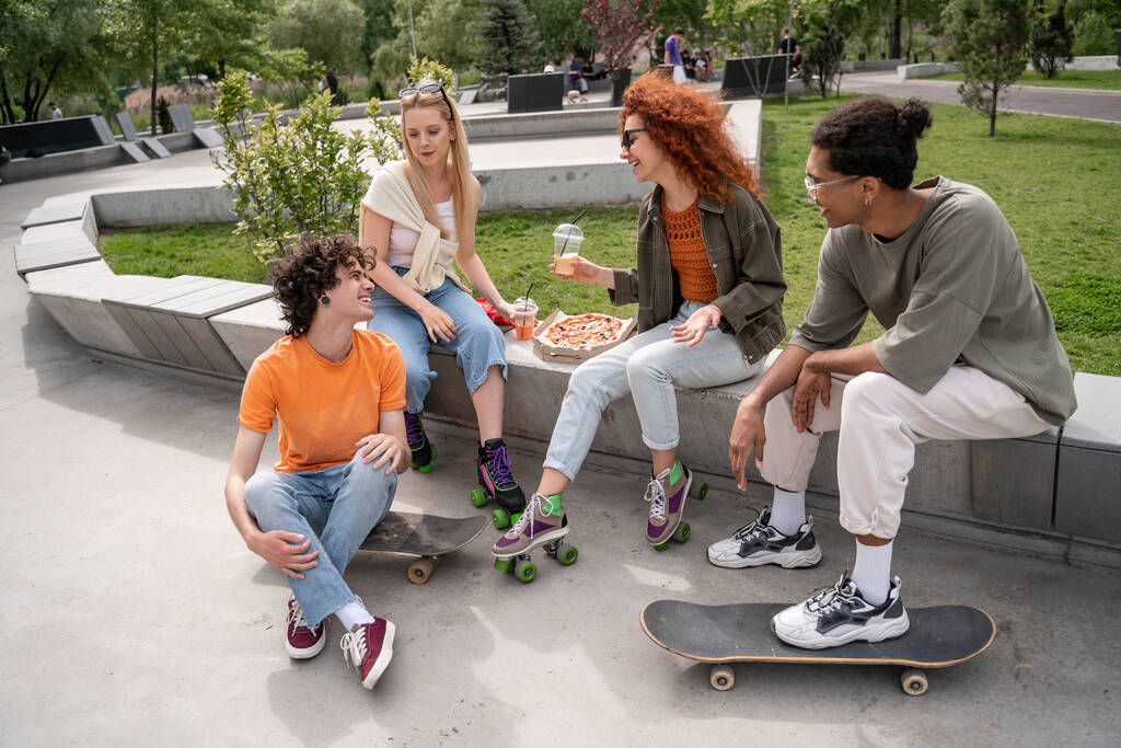 fröhliche multiethnische Skater unterhalten sich in der Nähe einer leckeren Pizza im Skatepark  - Foto, Bild