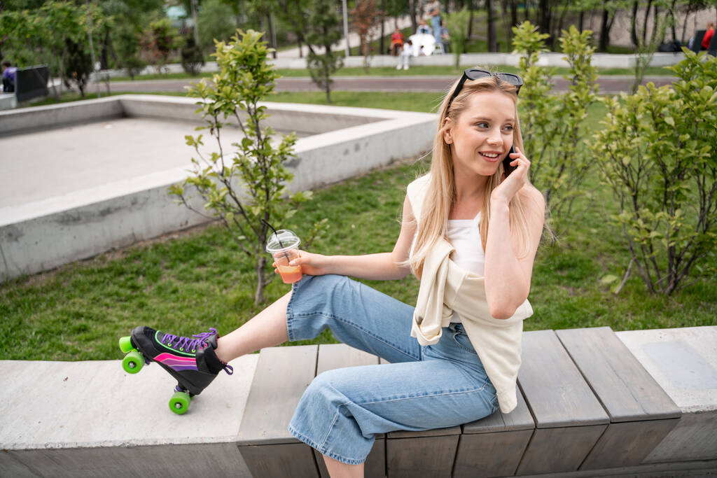 smiling roller skater with cocktail sitting on border bench and talking on mobile phone  - Photo, Image