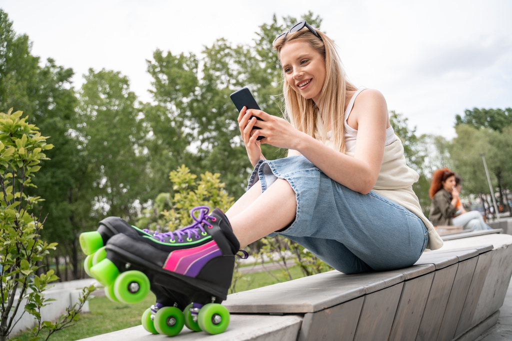 счастливая блондинка в роликах сидит на приграничной скамейке и отправляет сообщения по мобильному телефону - Фото, изображение