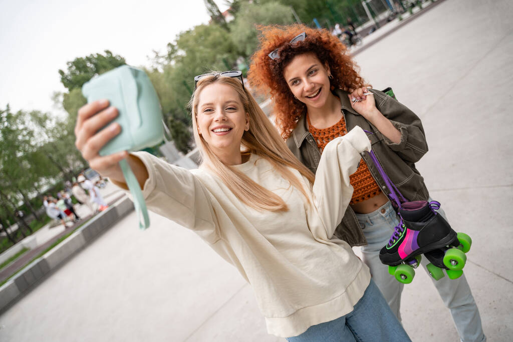 lockige Frau mit Rollschuhen lächelt neben Freund, der Selfie auf Digitalkamera macht - Foto, Bild