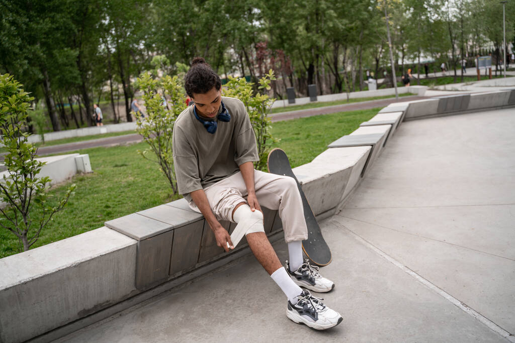 Genç Afrikalı Amerikalı kaykaycı bankta otururken dizini yaraladı. - Fotoğraf, Görsel