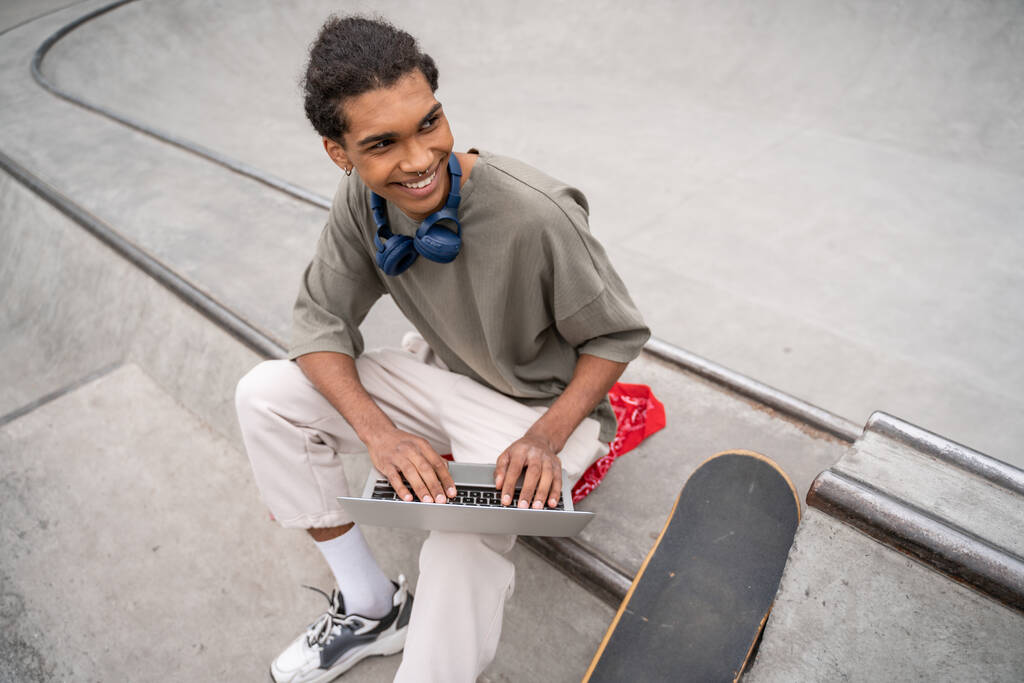 vista ad alto angolo di allegro studente afroamericano che utilizza il computer portatile sul confine vicino skateboard - Foto, immagini