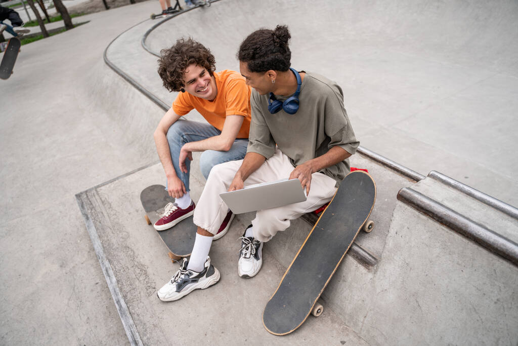feliz interracial skaters falando enquanto sentado no skate rampa com laptop - Foto, Imagem