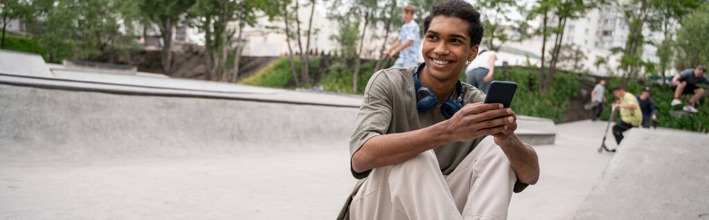 feliz homem americano africano com fones de ouvido e telefone celular sentado no parque de skate, banner - Foto, Imagem