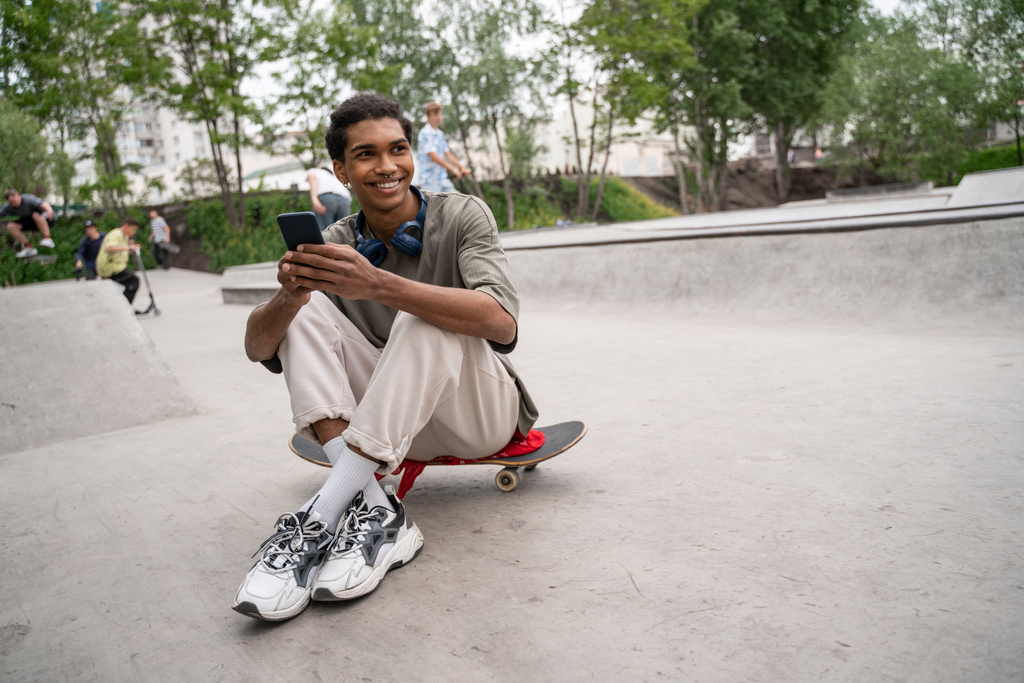 giovane e felice uomo africano americano utilizzando smartphone mentre seduto su skateboard - Foto, immagini
