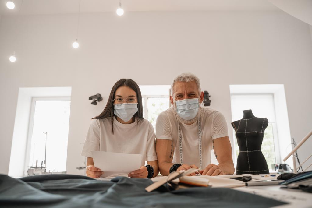 Mezirasoví návrháři v lékařských maskách při pohledu do kamery poblíž tkaniny  - Fotografie, Obrázek