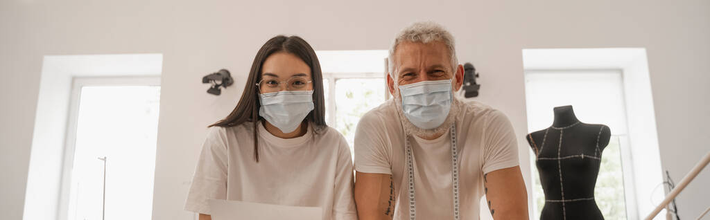 Межрасовые дизайнеры в медицинских масках смотрят в камеру в ателье, баннер  - Фото, изображение