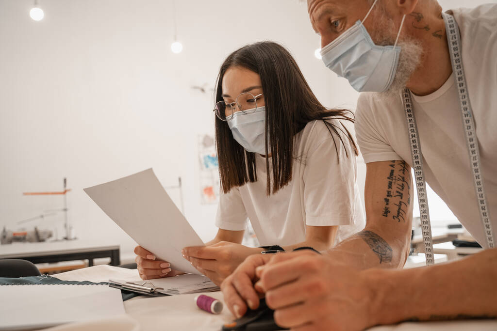 Tıp maskeli Asyalı tasarımcı atölyedeki meslektaşının yanında kağıt tutuyor.  - Fotoğraf, Görsel