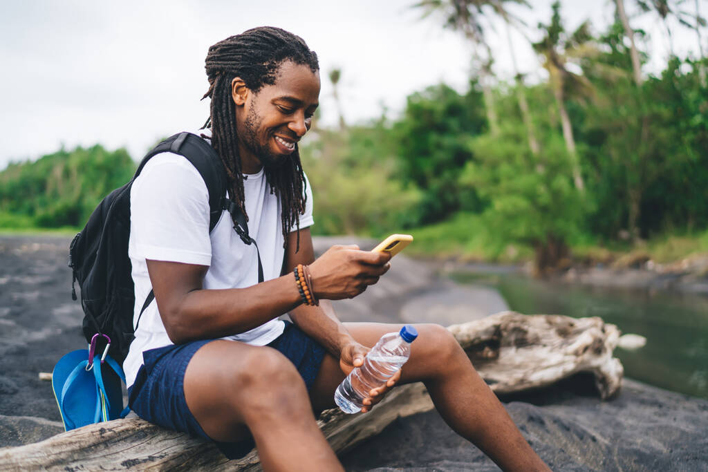Side view content African American male hiker sitting on dry tree trunk and browsing mobile phone while resting in tropical forest - 写真・画像