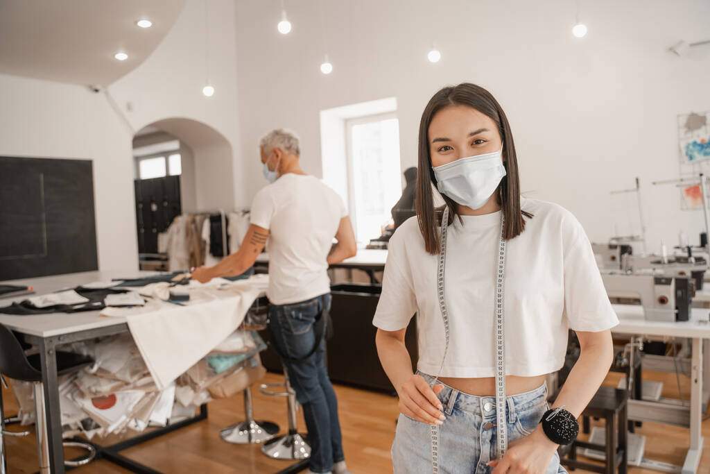 Ázsiai tervező orvosi maszk és mérőszalag a kamerába, miközben kolléga dolgozik a stúdióban  - Fotó, kép