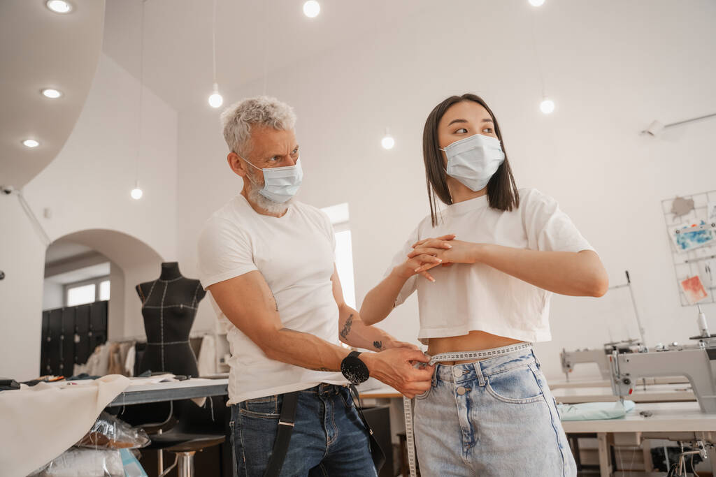 Designer in medical mask measuring waist of asian client in studio  - Photo, Image