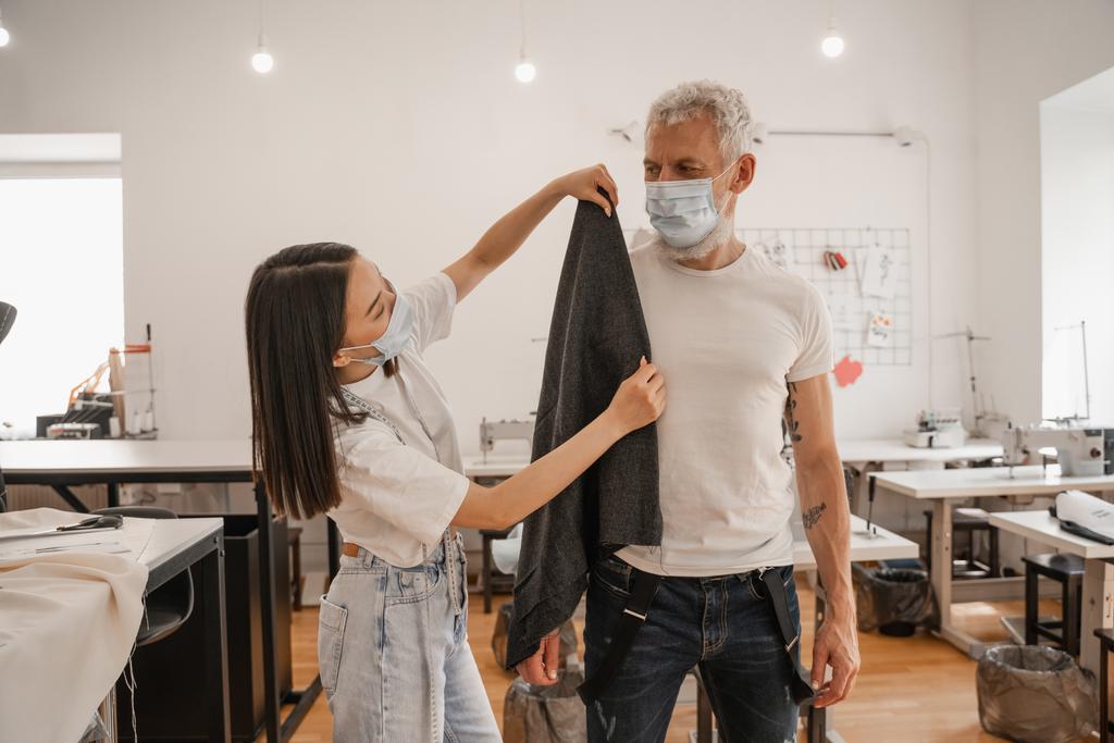Designer asiatique dans le masque médical tenant tissu près du client dans l'atelier  - Photo, image