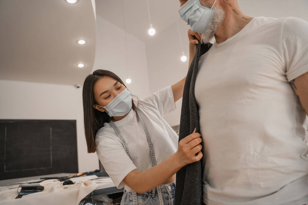 Asiatischer Designer mit medizinischer Maske und Maßband hält Tuch in Kundennähe  - Foto, Bild