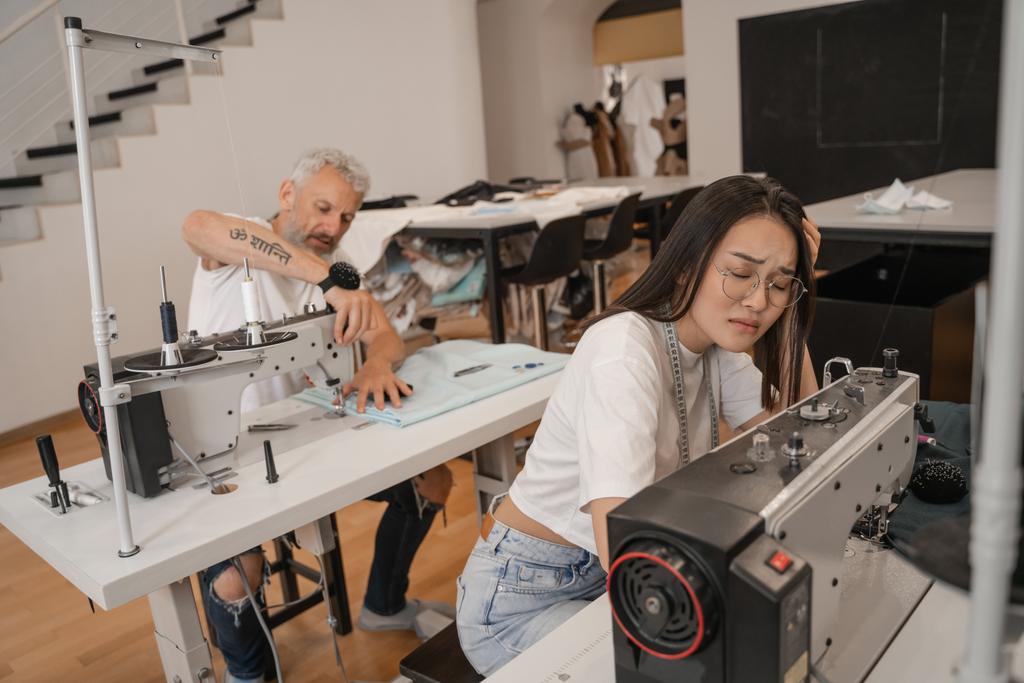 Tired asian seamstress sitting near sewing machine and colleague  - Photo, Image