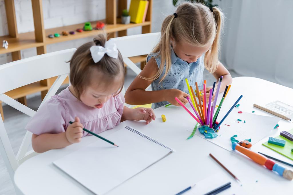Oyun odasındaki çocuğun yanında kağıt üzerine çizim yapan, Down sendromlu bir kızın yüksek açılı görüntüsü  - Fotoğraf, Görsel