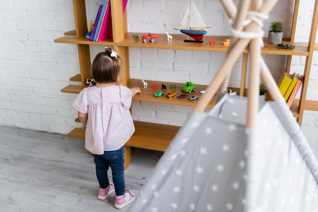 achteraanzicht van peuter meisje met down syndroom bereiken speelgoed op houten plank  - Foto, afbeelding